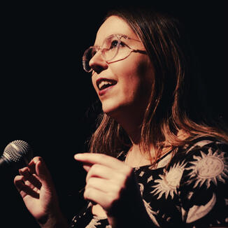 A photo of Ella, a white woman with brown hair and glasses. She's standing in a spotlight speaking into a microphone, looking up.